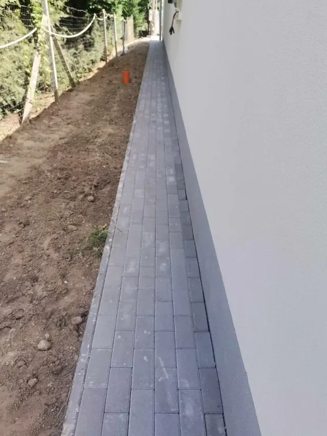 Sötétszürke téglakő járda a ház fala mentén, szegéllyel lezárva